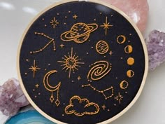 a black and gold embroidered hoop with stars, planets and clouds on it next to some crystals