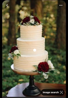 a wedding cake with red and white flowers on top is sitting on a table in the woods