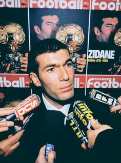 a man holding two soccer trophies in front of a wall covered with pictures and advertisements