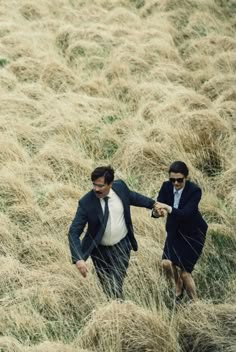 two men in suits are walking through tall grass near the water and one is holding his hand