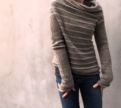 a woman standing in front of a concrete wall wearing a sweater and jeans with her hands on her hips