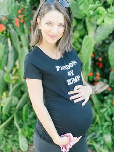 a pregnant woman wearing sunglasses standing in front of some plants and flowers with her hands on her belly