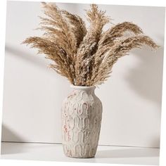 a white vase filled with dried grass on top of a table next to a wall