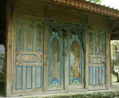 an old wooden building with blue and green paint on it's doors, in front of a tree