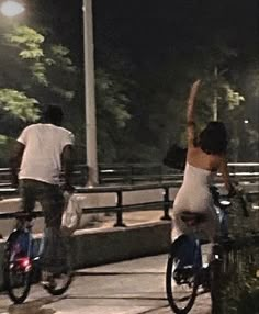 two people riding bikes down a sidewalk at night with their arms up in the air