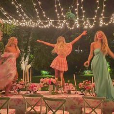 three women standing in front of a table with flowers on it and lights strung overhead