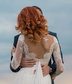 the back of a bride's dress as she stands next to her groom in front of an overcast sky