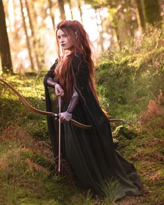 a woman with long red hair holding a bow and arrow in the woods, wearing a black dress