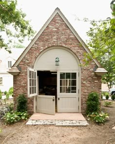 a small brick building with an open door and arched window on the front, surrounded by greenery