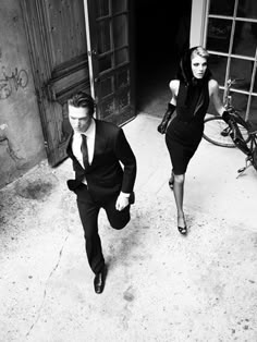 a man and woman are walking down the street in black suits, one is carrying a bike