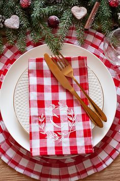 a red and white checkered table cloth with gold utensils on it, next to a christmas tree