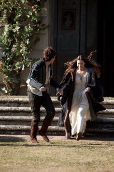 a man and woman dressed in period clothing walking on the grass with one holding the other's hand