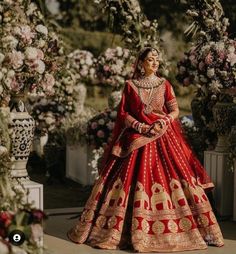 a woman in a red and gold bridal gown standing under an archway with flowers