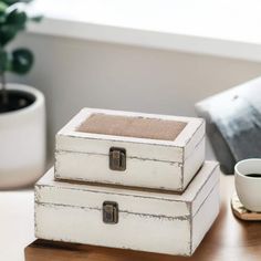 two white boxes sitting on top of a wooden table next to a cup and plant