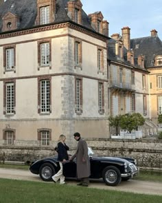 two people standing next to an old fashioned car in front of a large stone building