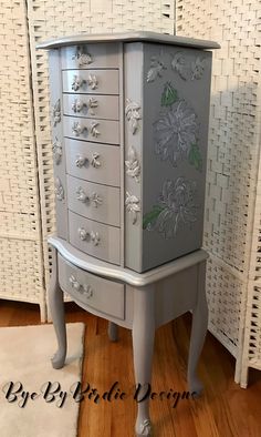 a white and grey cabinet with flowers painted on it's sides, in front of wicker wall