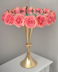 a gold vase filled with pink roses on top of a white table next to a wall