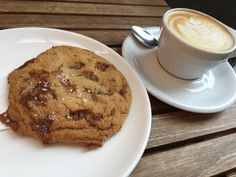 there is a chocolate chip cookie on the plate next to a cup of cappuccino