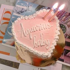 a pink heart shaped birthday cake with two lit candles on it and the words aquarius baby written in white frosting