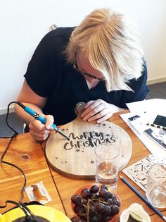 a woman sitting at a table cutting up a wooden sign with the words merry christmas written on it