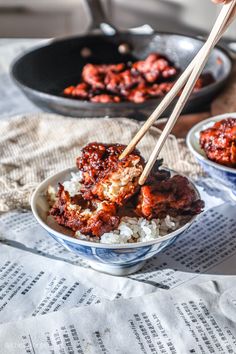 a person holding chopsticks over rice in a bowl with meat and sauce on the side