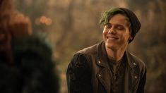 a young man with green hair smiles at the camera while wearing a leather jacket and beanie