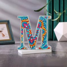 the letter m is made out of beads and sits on a table next to a framed photograph