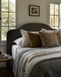 a bed with several pillows on it in front of two windows and a side table