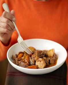 a person holding a fork in a bowl filled with meat and potatoes on a table