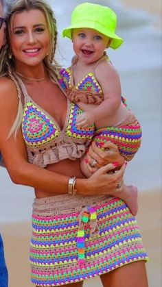 a woman holding a baby wearing a colorful bathing suit and yellow hat on the beach