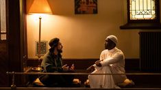 two men sitting on the floor in front of a window talking to each other and one is wearing a white turban