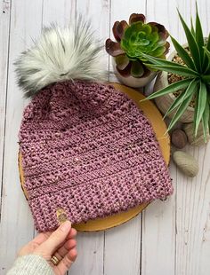 a hand is holding a knitted beanie on top of a wooden plate next to succulents