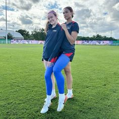 two girls standing on top of each other in front of a soccer field with their arms around one another