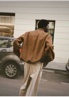 a man walking down the street in front of a building wearing a brown jacket and khaki pants