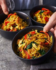 three bowls filled with noodles and vegetables on top of a table next to a person's hand