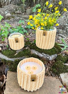 two knitted candle holders sitting on top of a moss covered ground with yellow flowers in the background