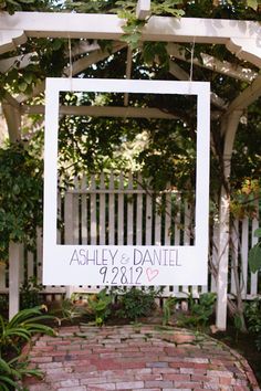 a wedding sign hanging from the side of a white arbor with greenery around it