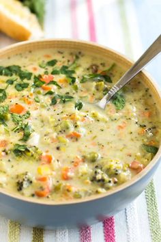 a bowl of soup with broccoli, carrots and bread in the background