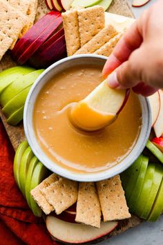 a person dipping an apple into a bowl of caramel sauce surrounded by apples and crackers