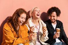 three people sitting next to each other and eating ice cream cones in front of a pink wall