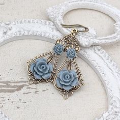 Shabby dusty blue-hued resin roses set upon vintage style brass filigree. I gave the roses a soft semi-matte finish. --------------------------------------------------------------------------------------------- Dimensions and details: - From the top of the lever-back ear wires to the bottom of the earrings, they measure approximately 2.3 inches (59mm) - Filigree is antiqued brass, made in the USA - Roses are made of resin - Ear wires are antiqued brass, lead-free and nickel-free, professionally finished in the USA --------------------------------------------------------------------------------------------- My other earrings may be found here: https://www.etsy.com/shop/ArdentHearts?section_id=12287411&ref=shopsection_leftnav_3 ---------------------------------------------------------------- Blue Ornate Handmade Earrings, Handmade Blue Ornate Earrings, Blue Rose Earrings, Elegant Rose Flower-shaped Earrings, Vintage Rose-colored Flower-shaped Jewelry, Rustic Barn Wedding, Resin Flowers, Rose Earrings, Dusty Blue