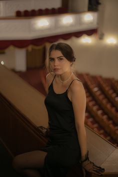 a woman in a black dress is sitting on the edge of a stage and looking at the camera