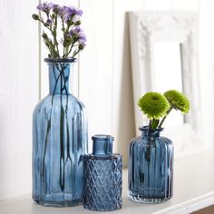 three blue vases with flowers in them on a white shelf next to a mirror