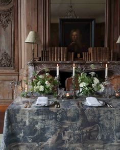 the table is set with candles, flowers and plates in front of an old fireplace