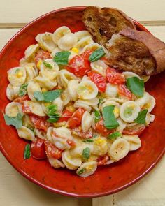 a red plate topped with pasta salad next to a piece of bread on top of a table