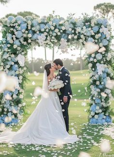 a bride and groom kissing in front of an archway with blue flowers on the grass