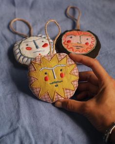 three handmade ornaments hanging from strings on a blue cloth covered bed with a person's hand holding one ornament in the foreground