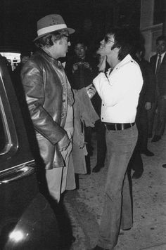 two men standing next to each other in front of a car and talking on the phone