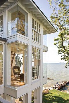 a white house sitting on top of a lush green field next to the ocean with lots of windows