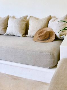 a white couch with pillows and a hat on it's back end, next to a plant